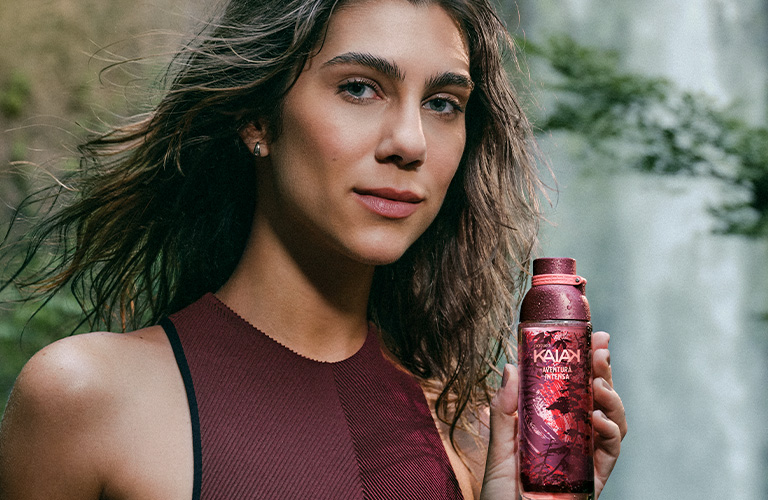 Una mujer con traje de baño rojo sostiene un frasco de perfume Kaiak rojo. Al fondo, una cascada y vegetación.
