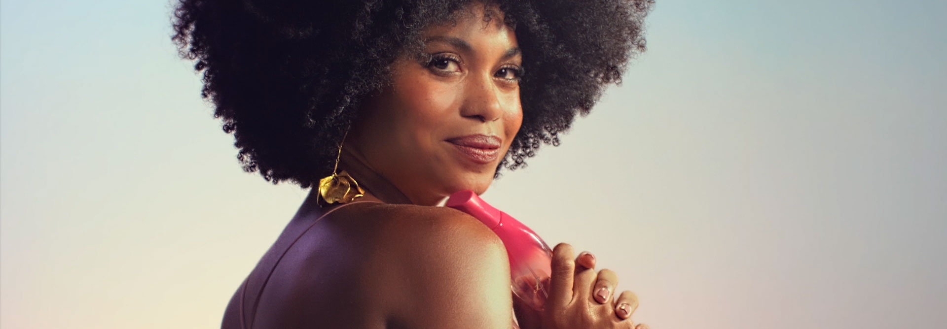Modelo negra de cabelos crespos usa vestido longo rosê com brincos e bracelete dourados. Na primeira cena, ela olha para a câmera e exibe a fragrância Natura Luna Divina. Na sequência, ela borrifa a fragrância no pescoço e no colo. Nas cenas seguintes, ela rodopia suavemente e sorri para a câmera segurando Natura Luna Divina nas mãos.