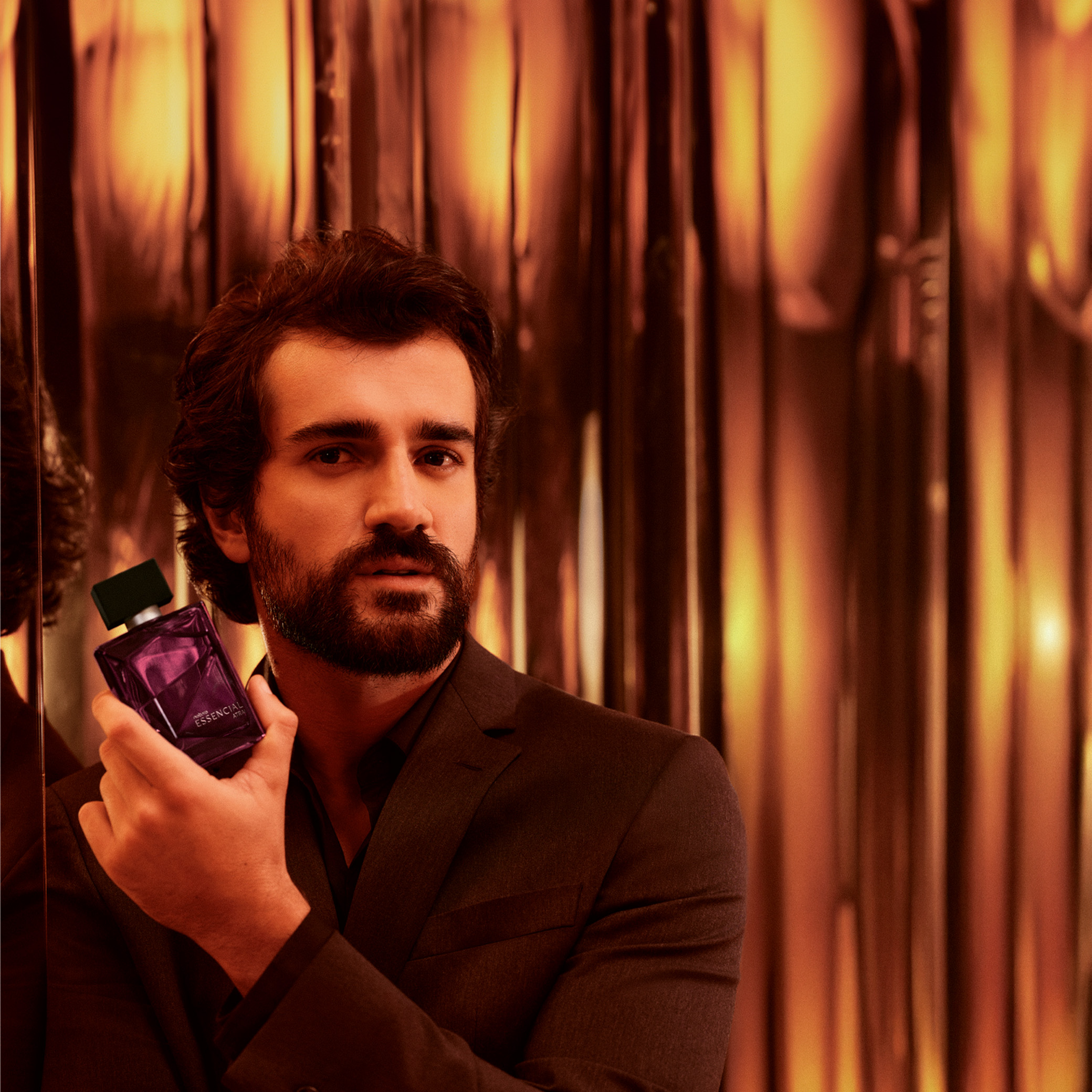 Homem de cabelo castanho e barba segura o Deo Parfum Essencial Atrai ao lado do rosto. A embalagem é na cor vinho, o modelo usa terno preto e o ambiente tem luzes quentes e tons dourados. 