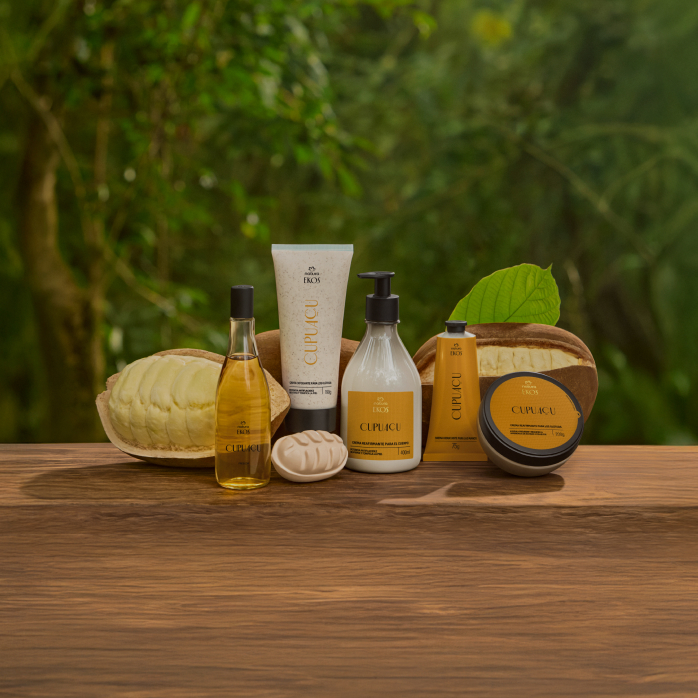 6 produtos de Ekos Cupuaçu, incluindo Frescor, Esfoliante, Sabonete e Cremes para Corpo e Mãos. Embalagens com tampas pretas e rótulos amarelos sobre superfície de madeira com plantas ao fundo.