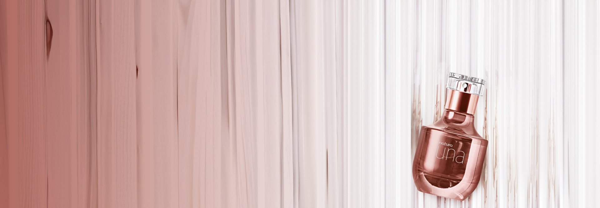 Frasco na cor rosê gold com tampa transparente do Deo Parfum Una. O produto está inclinado sobre uma superfície de madeira com tons rosados.