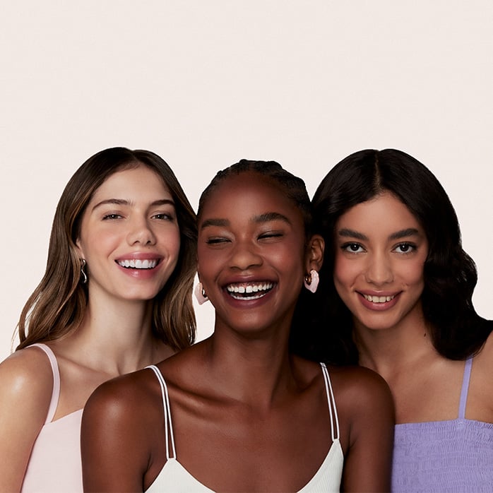 Três mulheres jovens com diferentes tons de pele sorriem juntas. Elas vestem blusas de alças finas em tons pastéis contra um fundo neutro claro. A mulher ao centro ri alegremente, enquanto as outras duas sorriem suavemente para a câmera.