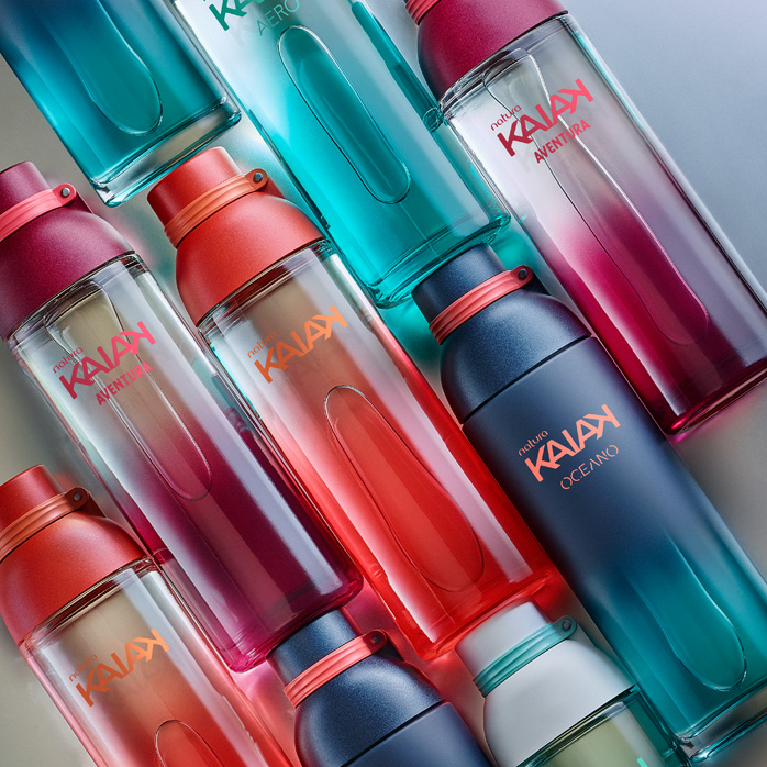 Frascos de diferentes fragrâncias Kaiak, em rosa, vermelho, azul e verde. Itens em diagonal sobre um fundo prateado.