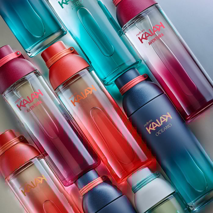 Frascos de diferentes fragrâncias Kaiak, em rosa, vermelho, azul e verde. Itens em diagonal sobre um fundo prateado.
