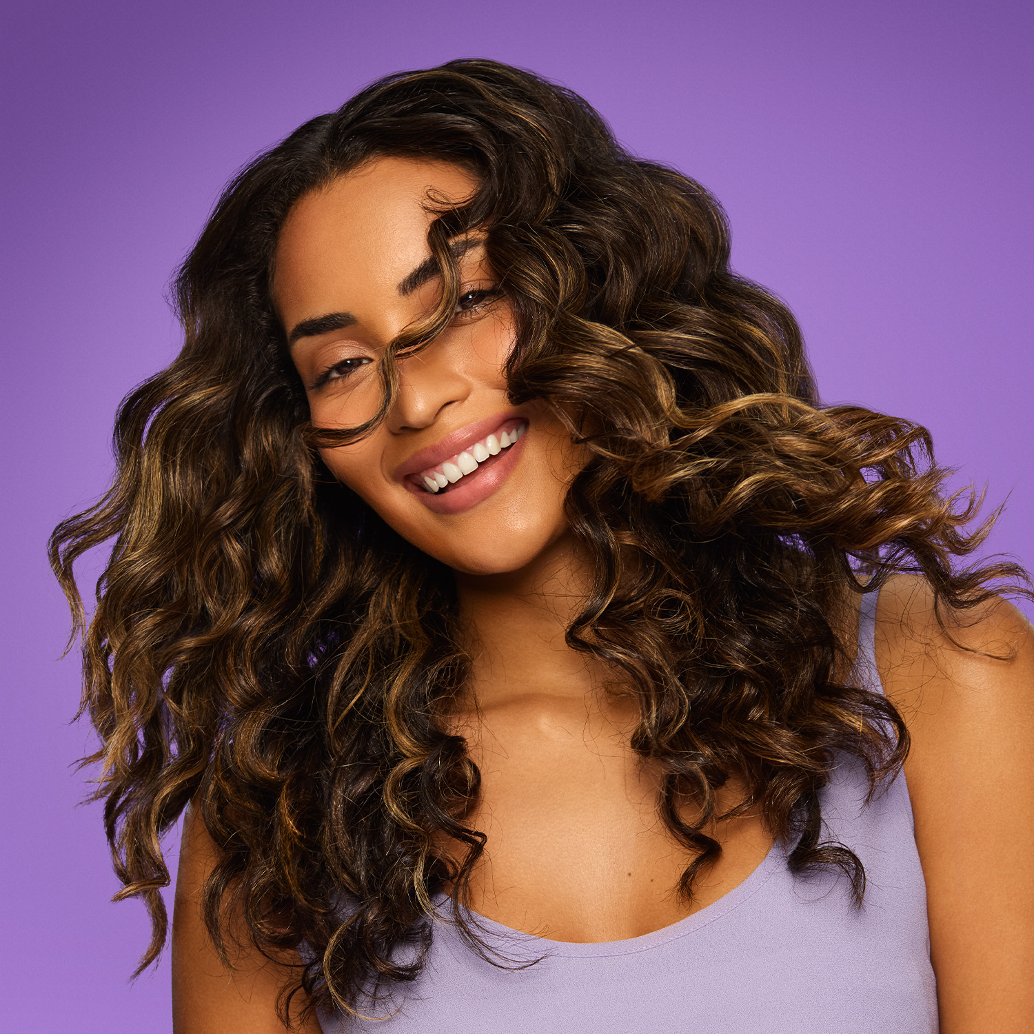 Mulher jovem de pele bronzeada sorri enquanto joga o cabelo. Seu cabelo é cacheado com ondas largas, na cor castanho com luzes douradas, com muito movimento e brilho.
