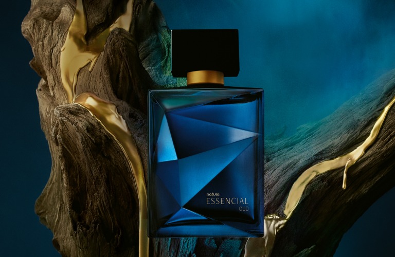 Perfume doce Essencial Oud Masculino em frente a um tronco de árvore. O fundo da imagem é azul, assim como o vidro