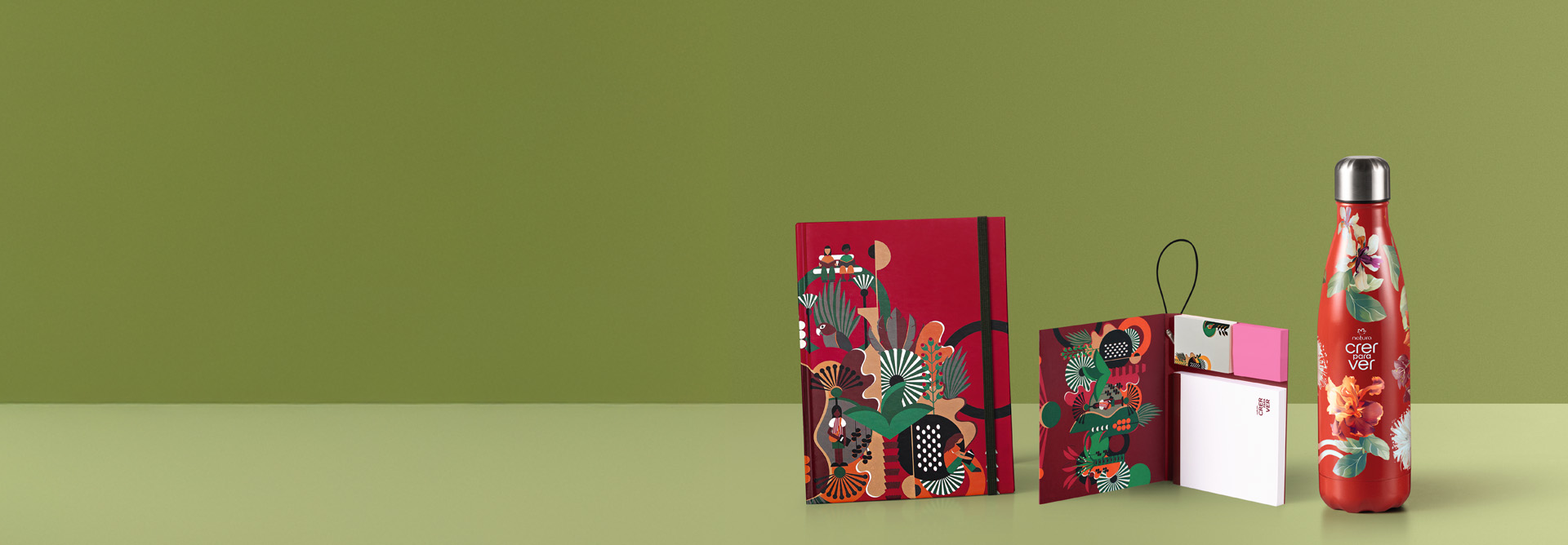 Itens da linha Natura Crer Para Ver, agenda vermelha estampada, bloco de notas aberto com notas brancas e cor-de-rosa e garrafa vermelha com tampa prateada e estampa floral. Fundo verde.