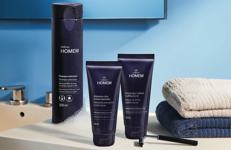 Foto de produtos Natura Homem sobre bancada de banheiro, com fundo azul, toalhas e aparelho de barbear ao lado.