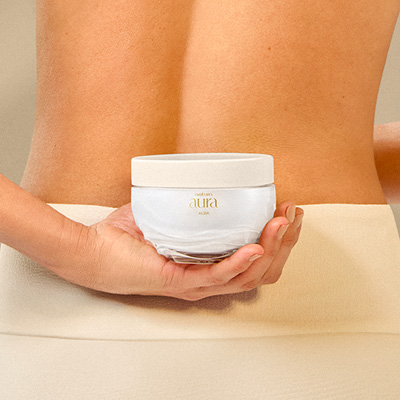 Mulher de pele bronzeada segura o Creme Corporal Aura Alba nas costas. A embalagem é um pote branco com tampa dourada.