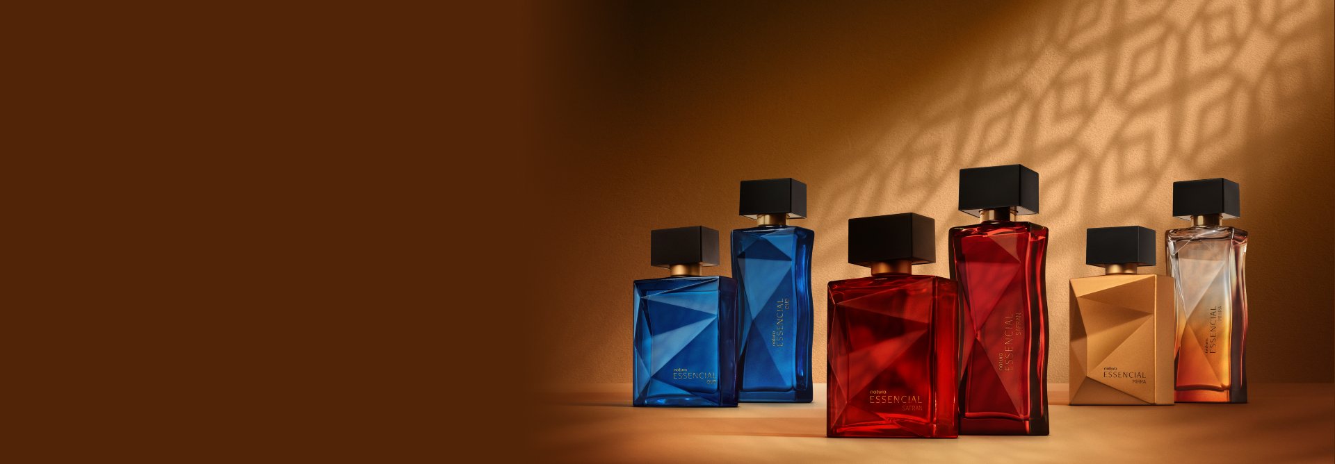 6 frascos de fragrâncias sendo 2 azuis de Essencial Oud, 2 vermelhos de Essencial Safran e 2 alaranjados de Essencial Mirra, todos com tampas pretas. Estão em fundo marrom com sombra geométrica.