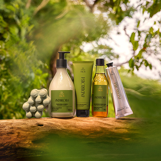 Kit Natura Ekos Andiroba com cinco produtos: sabonete massageador, esfoliante térmico, creme, óleo e polpa para mãos em embalagens verdes e bege, dispostos sobre tronco em meio à natureza.