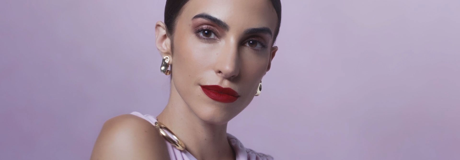 Close de uma mulher branca com cabelos castanhos-escuros presos em um coque, batom vermelho e sombra avermelhada, usando brincos e colar dourados. Ela veste uma blusa lilás sobre fundo lilás.
