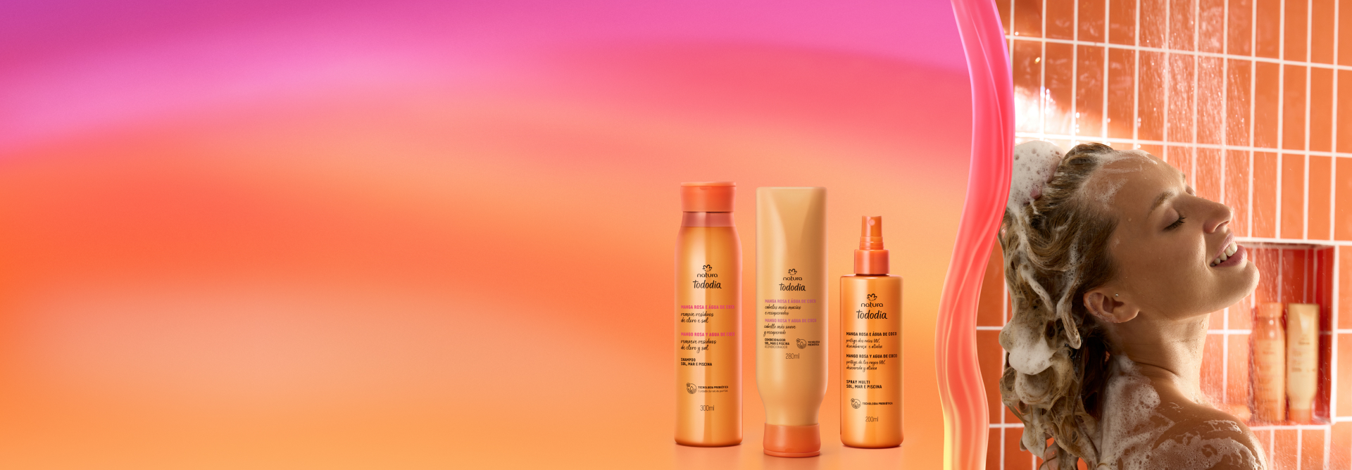 Uma mulher sorridente e de cabelos longos, tomando banho sob um chuveiro com azulejos de cor tangerina, ensaboando a cabeça com espuma abundante. Em primeiro plano, três produtos de cabelo da linha Tododia Verão, da Natura, com embalagens em tons de laranja e bege, estão dispostos sobre um fundo degradê em cores quentes (rosa, laranja e amarelo). Os produtos são um shampoo, um condicionador e um spray.