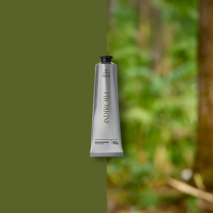 Bisnaga prata de Fluido de Massagem para o Corpo Ekos Andiroba, em fundo de cenário de floresta desfocada à direita e verde à esquerda.