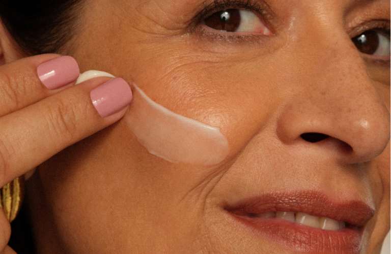 Mulher aplicando um creme na região do olho com a ponta dos dedos, close-up destacando linhas de expressão e unhas rosa.
