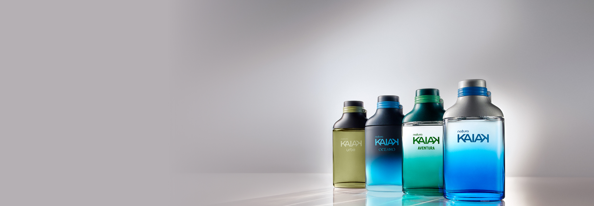 Frascos de fragrâncias da linha Natura Kaiak, nas cores verde, azul-escuro, verde-claro e azul-claro, em bancada reflexiva e fundo cinza.