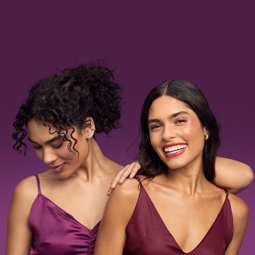 Foto de duas mulheres, uma de cabelos cacheados e presos e outra com os cabelos soltos, em fundo roxo. Ambas usam vestido de alças em tons de vinho.