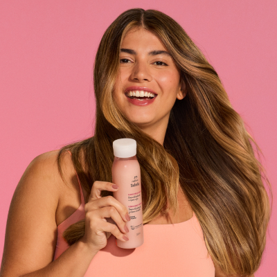 Mulher de cabelos longos e lisos com mechas loiras, sorrindo enquanto segura um shampoo Natura Tododia Flor de Cereja e Abacate (embalagem rosa-claro) ao lado de seu corpo. O foco está no cabelo com brilho e no produto. Fundo rosa.