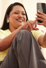Mulher de pele morena e cabelos longos e escuros, usa blusa amarela e calça cinza. Ela está sentada em uma poltrona caqui e sorri, enquanto mexe no celular sorrindo.