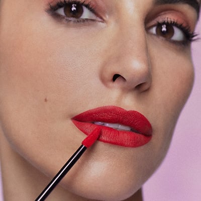 Close-up no rosto de uma mulher branca que está aplicando batom vermelho vibrante com um aplicador preto, sobre um fundo roxo-claro e desfocado. 