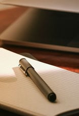 Close-up de uma caneta escura deitada sobre um caderno de papel quadriculado em ângulo diagonal.