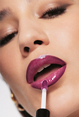Mulher de pele branca aplica um gloss roxo brilhante sobre os lábios delineados em tom magenta. Nos olhos, ela usa maquiagem esfumada em tons de marrom.