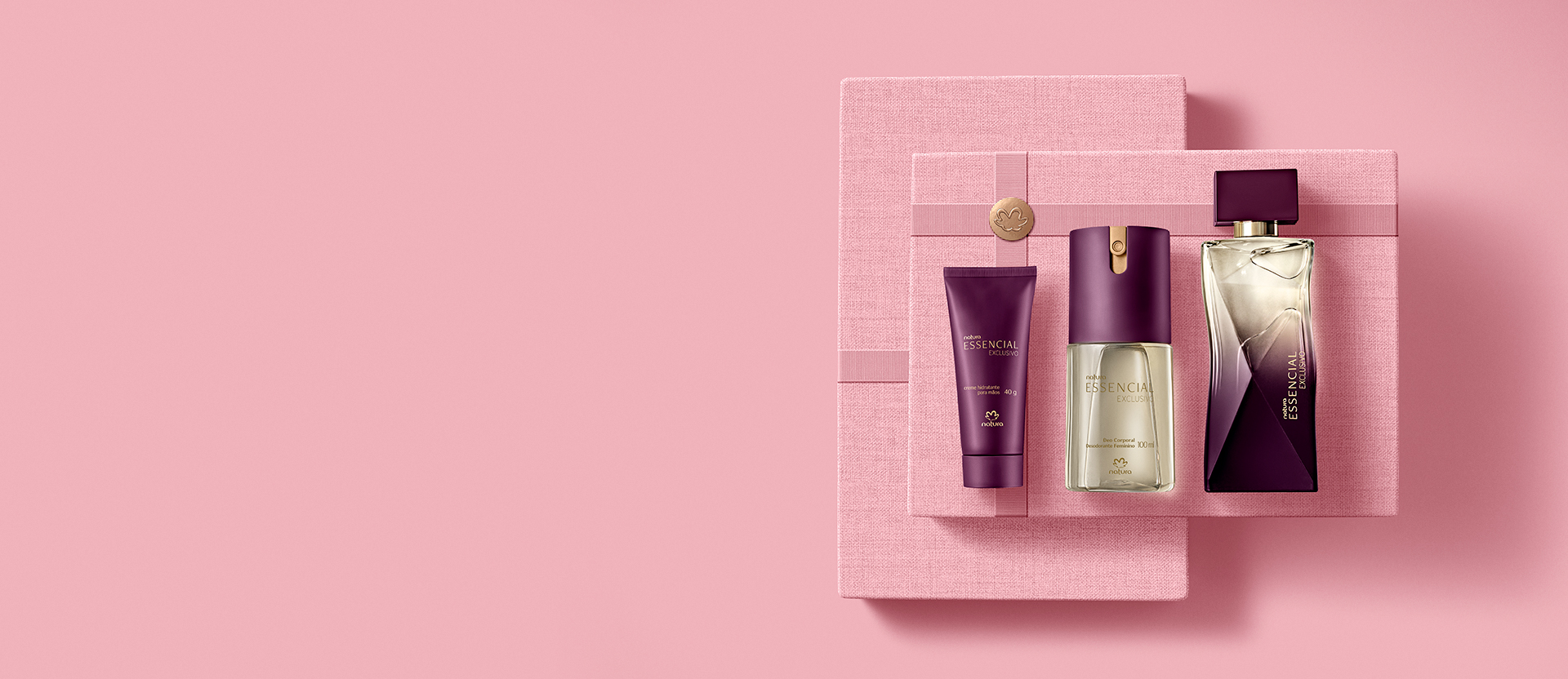 Kit de produtos sobre caixas rosas texturizadas. O kit contém um perfume, um creme hidratante e um óleo corporal.