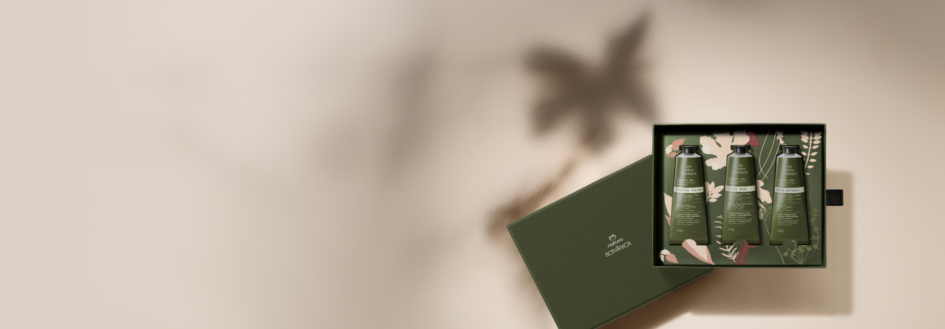 Três bisnagas dos novos Cremes para as Mãos Natura Bothânica, com embalagens verde-escuro e tampas pretas, destacam-se em caixa decorada de mesmo tom. Fundo bege com folhagens em efeito desfocado.
