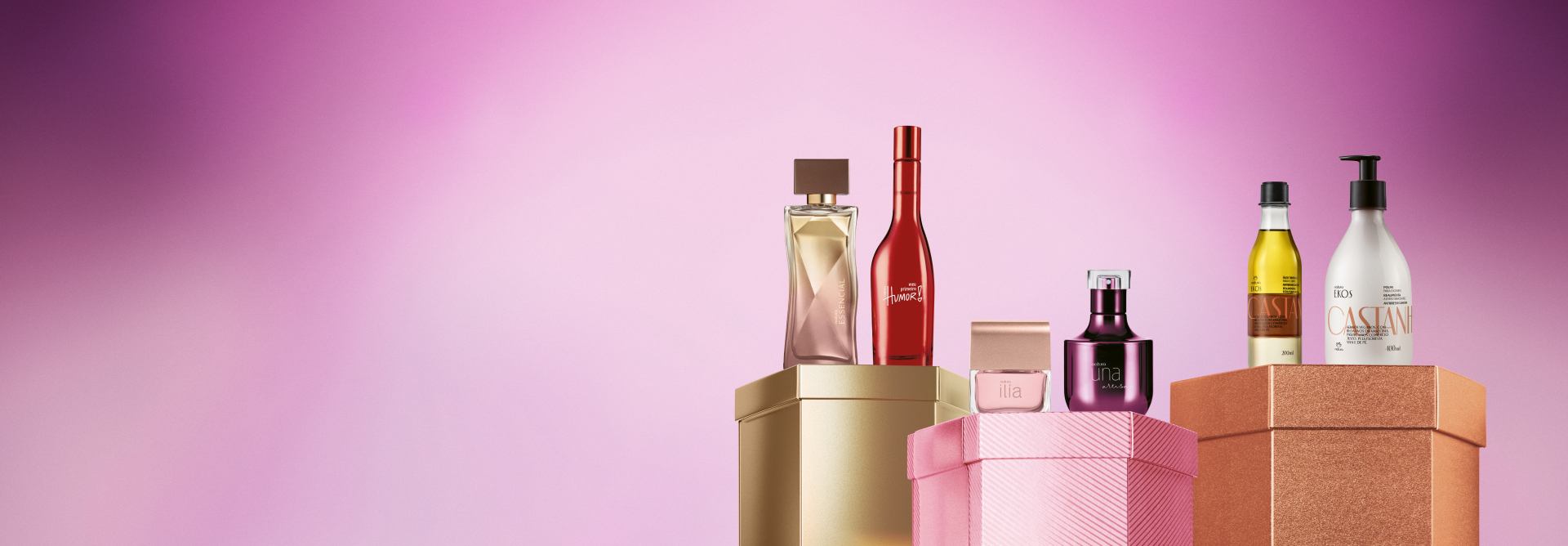 Embalagens Natura sobre caixas brilhantes dourada, rosa e laranja, em fundo rosa: frascos das fragrâncias Essencial, Meu Primeiro Humor, Ilía, Una Artisan e de óleo e polpa para o corpo Ekos Castanha.
