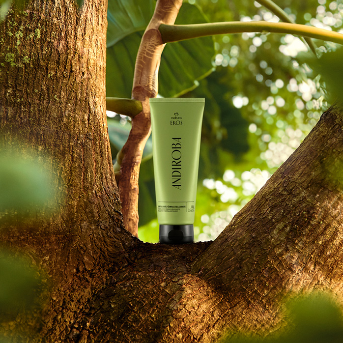 Bisnaga verde com tampa preta de Esfoliante Térmico Relaxante Ekos Andiroba sobre tronco de árvore, em cenário de floresta desfocada.