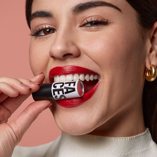 Close do rosto de uma mulher sorrindo e segurando um batom da linha Natura Faces entre os dentes. Ela usa um batom vermelho vibrante.