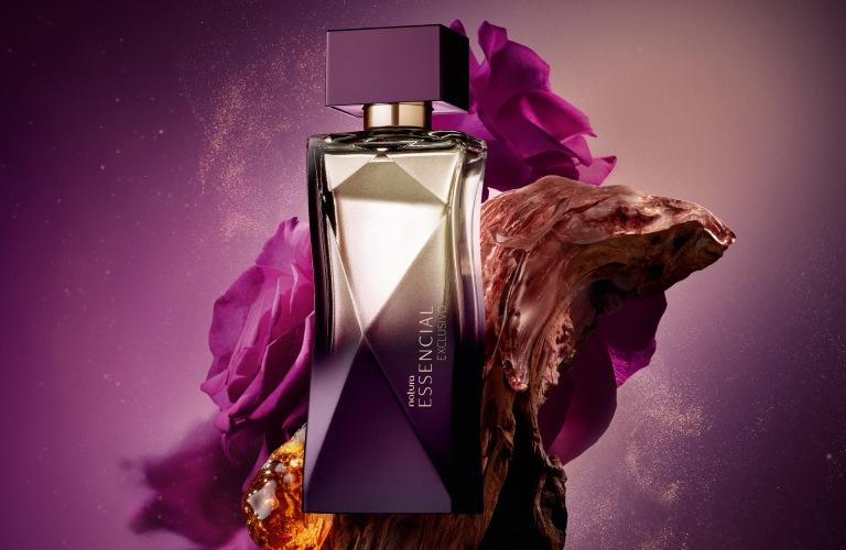 Perfume doce Essencial Exclusivo feminino posicionado na frente de um pedaço de madeira. O vidro é uma mescla de roxo e amarelo