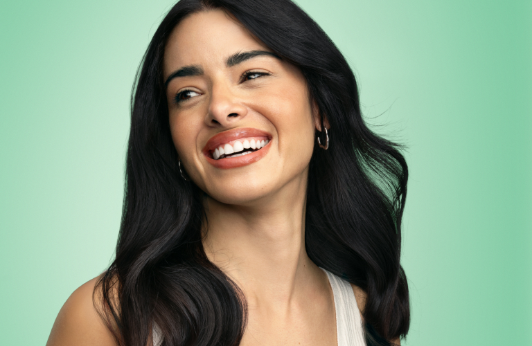 Foto de mulher sorrindo, com fundo verde liso. Ela usa top branco e tem os cabelos longos e soltos sobre os ombros.