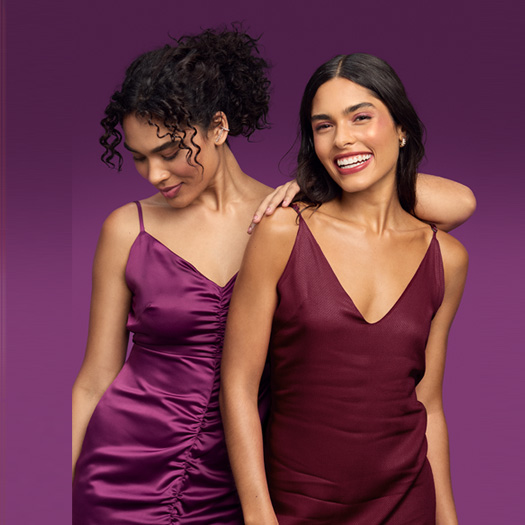 Foto de duas mulheres, uma de cabelos cacheados e presos e outra com os cabelos soltos, em fundo roxo. Ambas usam vestido de alças em tons de vinho.