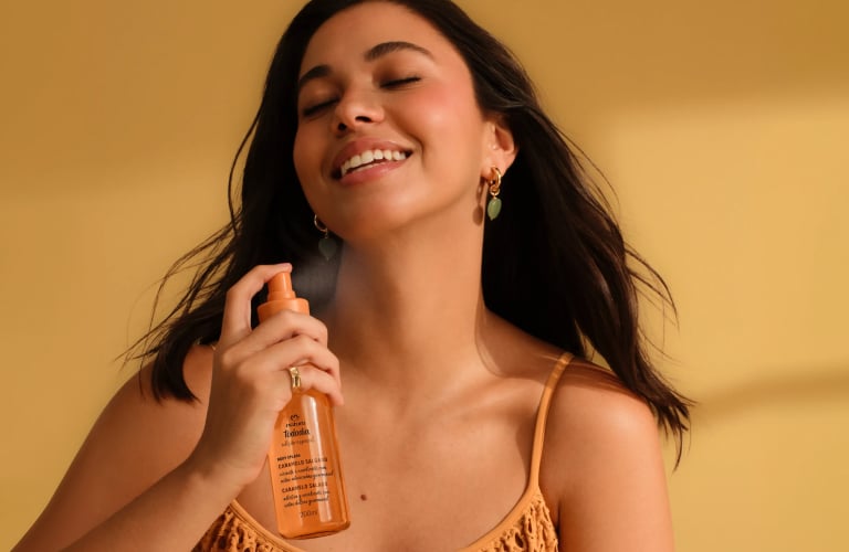 Foto de mulher sorrindo sobre fundo amarelo, borrifando body splash Natura Tododia no pescoço, com névoa do produto visível no ar.