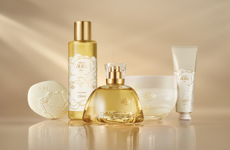 Foto. Fundo bege com luz suave. Cinco produtos da linha Natura Aura Alba: frasco spray, perfume, hidratante, sabonete e creme. Embalagens brancas e douradas.