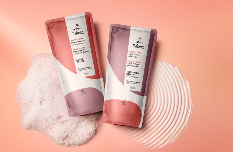 Foto com fundo em degradê rosa, dois refis Natura Tododia de shampoo e condicionador apoiados sobre espuma e textura circular branca.