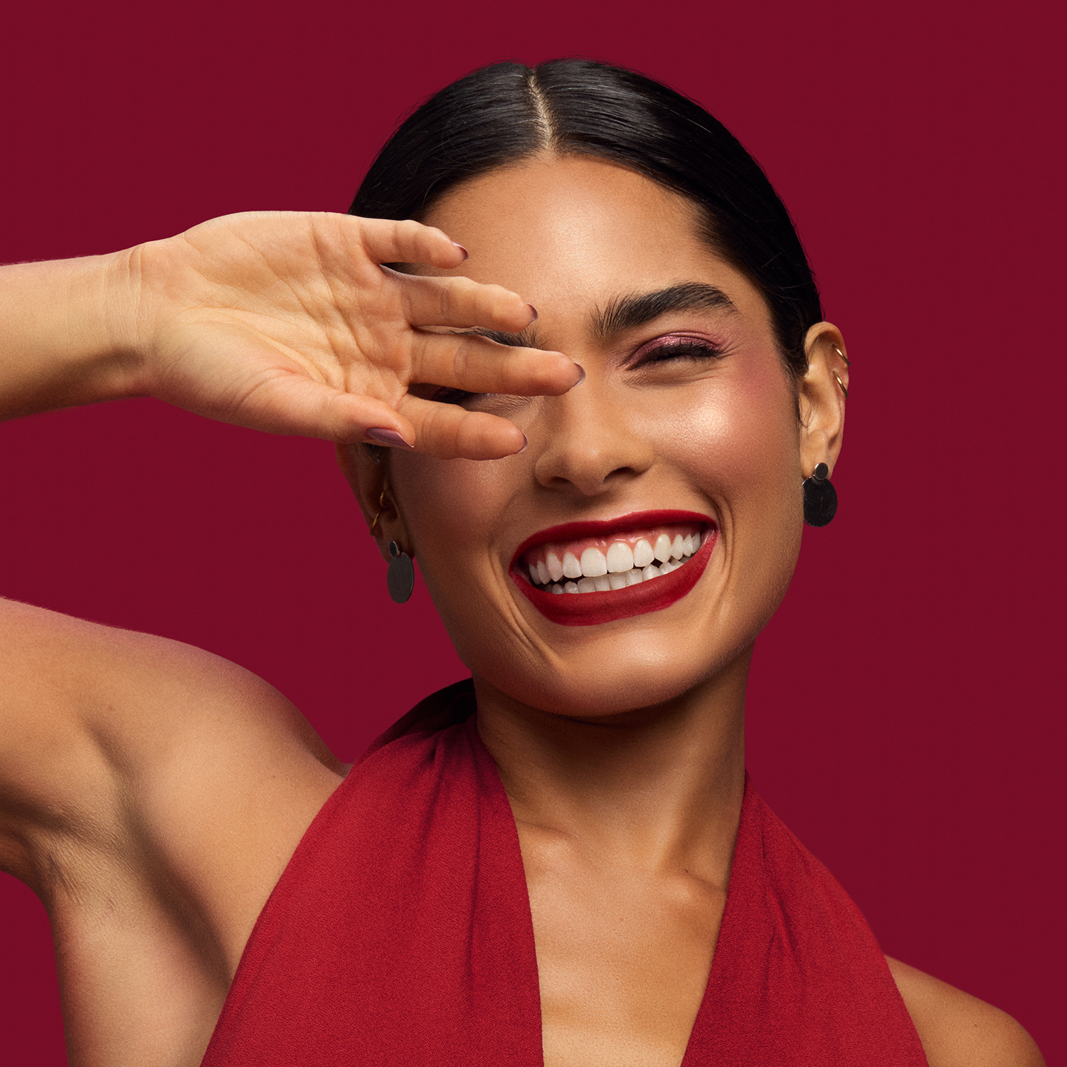 Mulher jovem de pele bronzeada e cabelo preto preso sorri enquanto leva a mão ao rosto. Ela usa um vestido vermelho e batom vermelho vibrante no tom cereja. Fundo vermelho. 