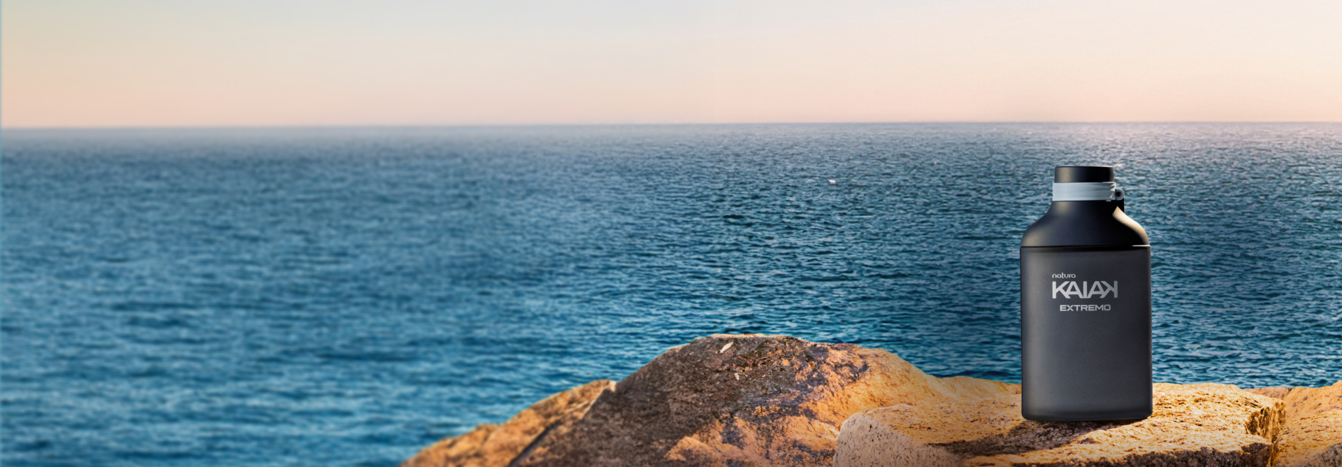 Desodorante Colônia Kaiak Extremo Masculino sobre uma rocha amarela e fundo com oceano azul e céu claro. Embalagem em azul marinho com detalhes em azul claro.