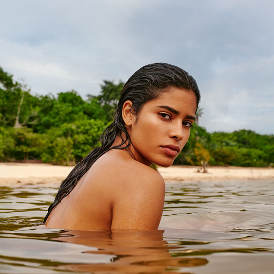 Mulher de pele morena e cabelo escuro molhado está parcialmente submersa em rio, olhando para trás. Ao fundo, há vegetação verde e uma faixa de areia sob céu nublado.