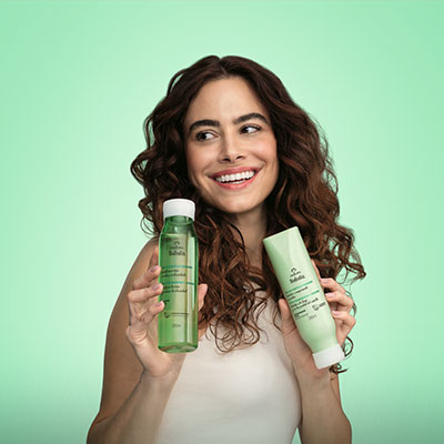 Mulher de cabelos longos e lisos com mechas loiras, sorrindo enquanto segura um shampoo Natura Tododia Flor de Cereja e Abacate (embalagem rosa-claro) ao lado de seu corpo. O foco está no cabelo com brilho e no produto. Fundo rosa.