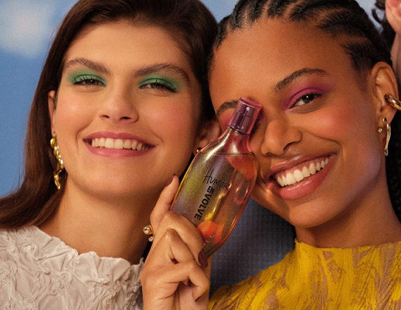 Duas mulheres olham para a câmera. À esquerda, mulher de pele clara e cabelos castanhos com blusa branca e, à direita, mulher de pele negra e tranças segura uma fragrância Natura Humor Envolve.