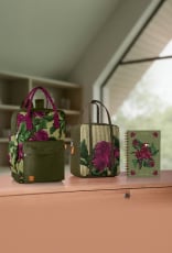 Mochila, frasqueira térmica e caderno Crer Para Ver, todos em tons de verde com estampas de folhagens e flores em tons de roxo e branco, repousam sobre uma bancada de madeira de uma sala com janelas.