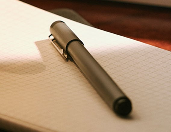 Close-up de uma caneta escura deitada sobre um caderno de papel quadriculado em ângulo diagonal.