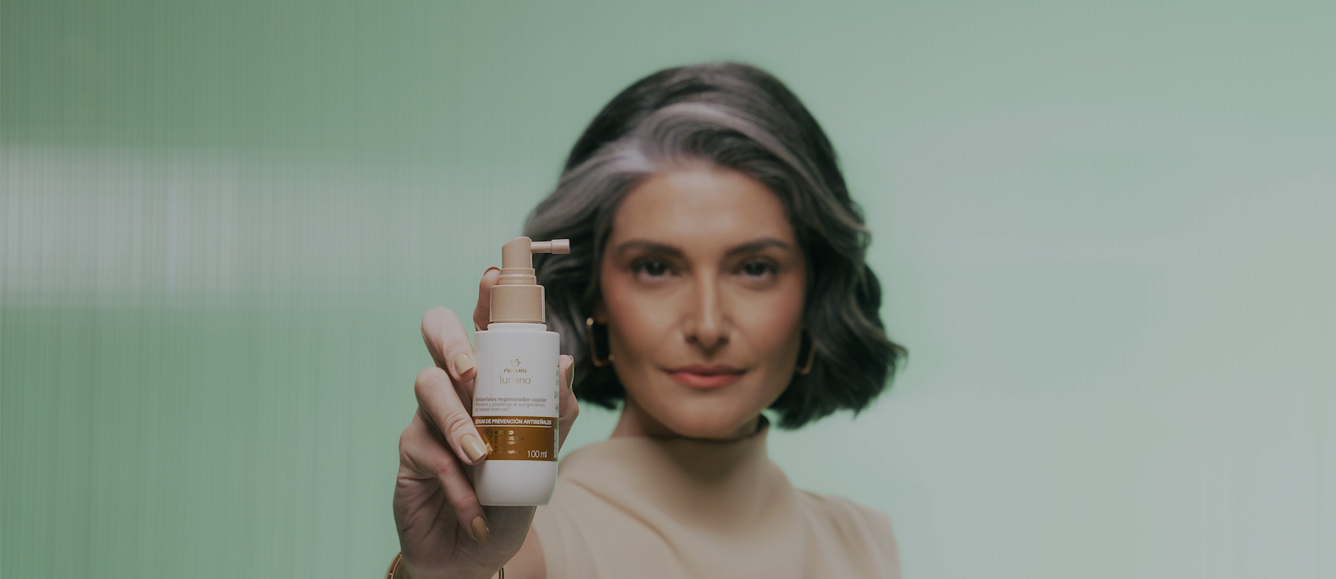 Vídeo com sequência de imagens: mulher branca de cabelos médios e grisalhos usando blusa bege, imagem de produtos da linha Natura Lumina Antissinais, mulher aplicando itens nos cabelos, animação de gota dourada e sua ação no couro cabeludo e fios de cabelo. Vídeo finaliza com mulher segurando item próximo ao corpo, em fundo verde.