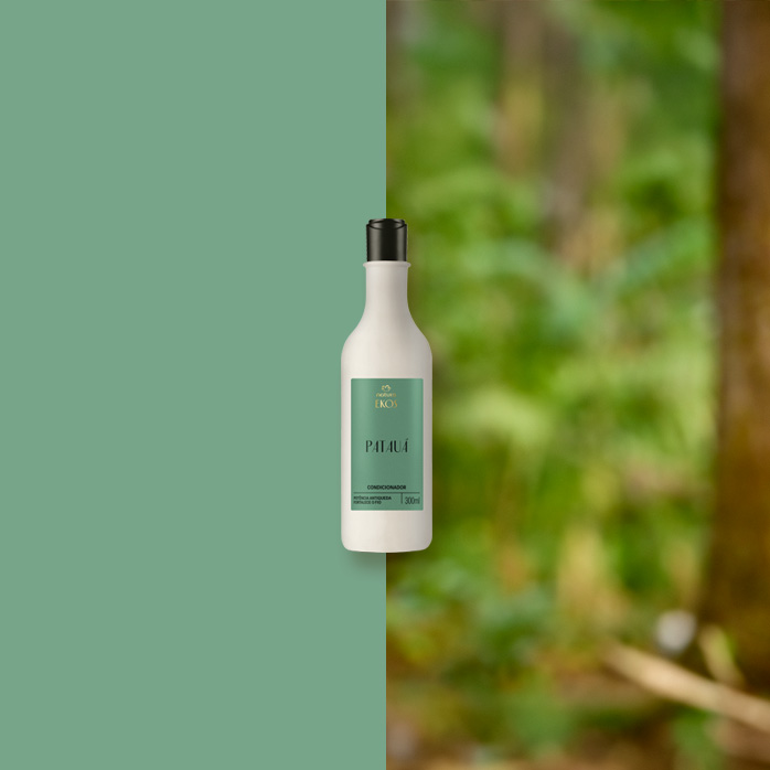Frasco branco de Condicionador Ekos Patauá com rótulo verde e tampa preta está centralizado, dividido entre fundo verde sólido e floresta desfocada ao fundo.