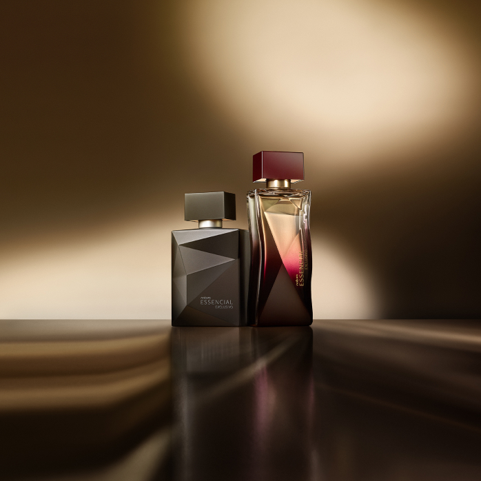 Essencial Exclusivo Feminino e Masculino, com frascos em tons de preto e violeta, aparecem iluminados sobre superfície marrom-acinzentado e fundo amarronzado.