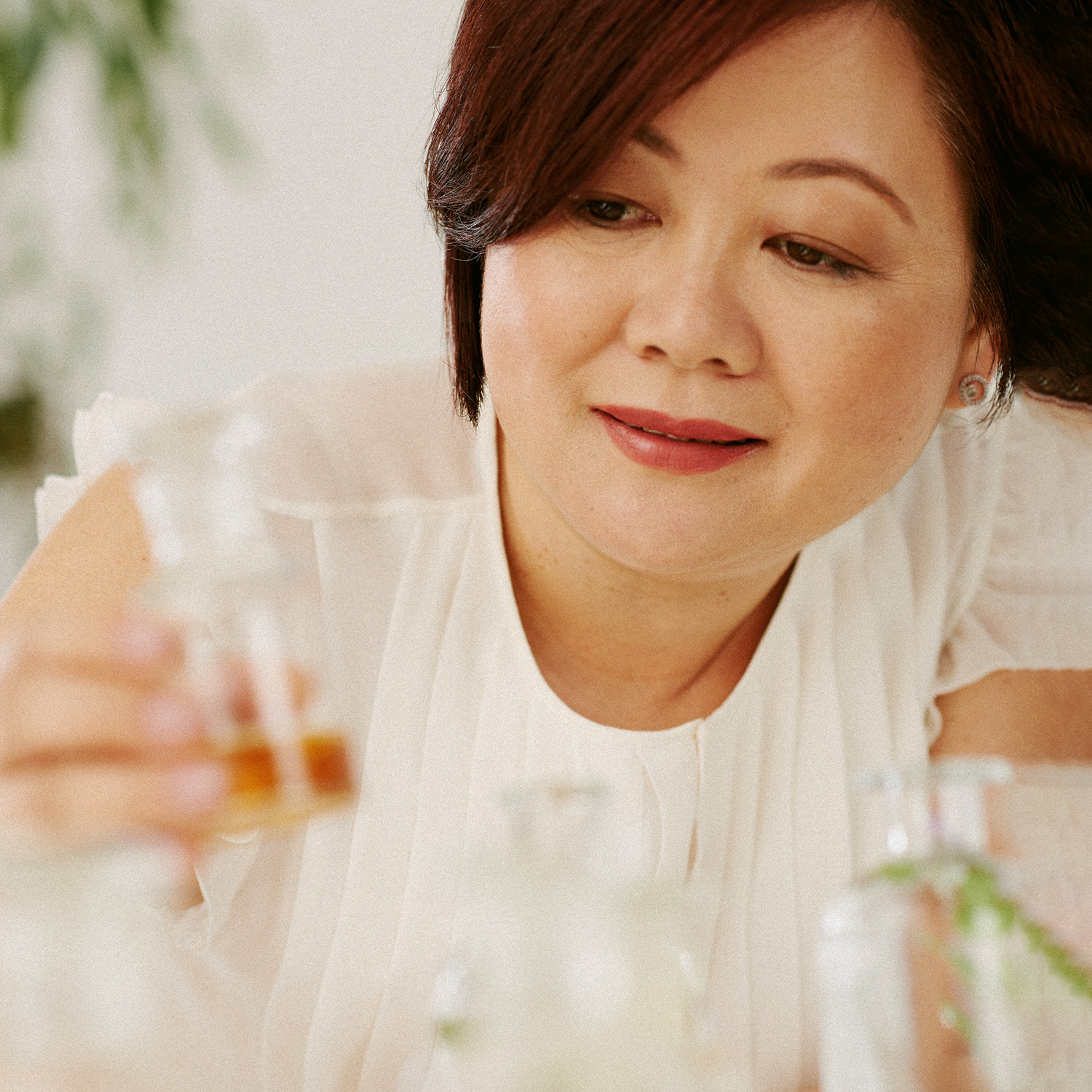 A perfumista Verônica Kato segura um frasco de vidro com essência. Outros frascos com ingredientes estão em cena desfocados. 