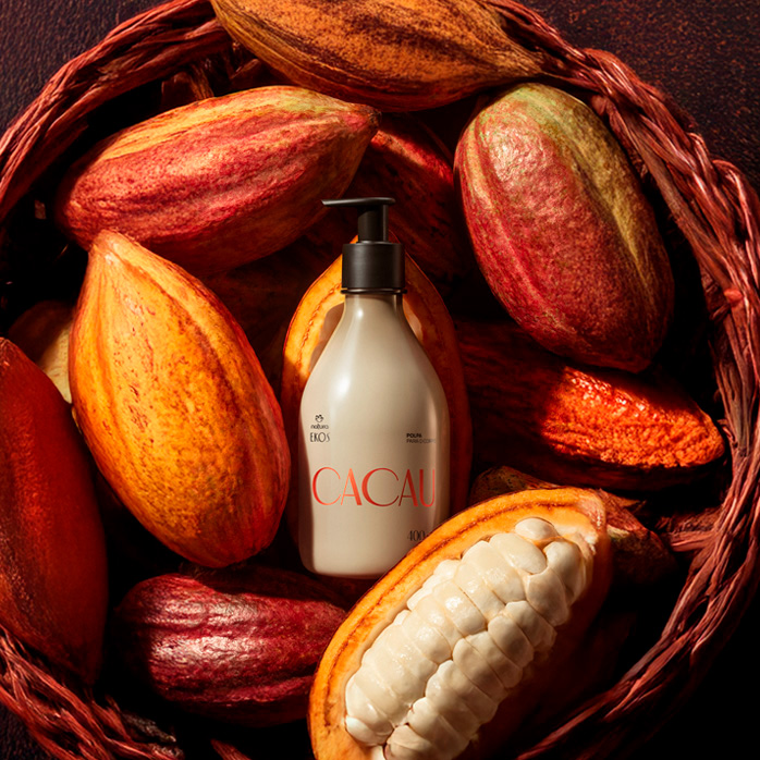  Frasco bege com pump preto de Polpa Desodorante Hidratante Corporal Ekos Cacau 400 ml está entre frutos de cacau inteiros e abertos, dentro de um cesto marrom em fundo escuro.