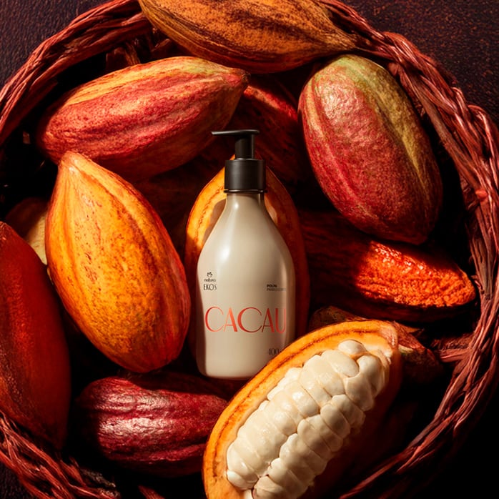  Frasco bege com pump preto de Polpa Desodorante Hidratante Corporal Ekos Cacau 400 ml está entre frutos de cacau inteiros e abertos, dentro de um cesto marrom em fundo escuro.